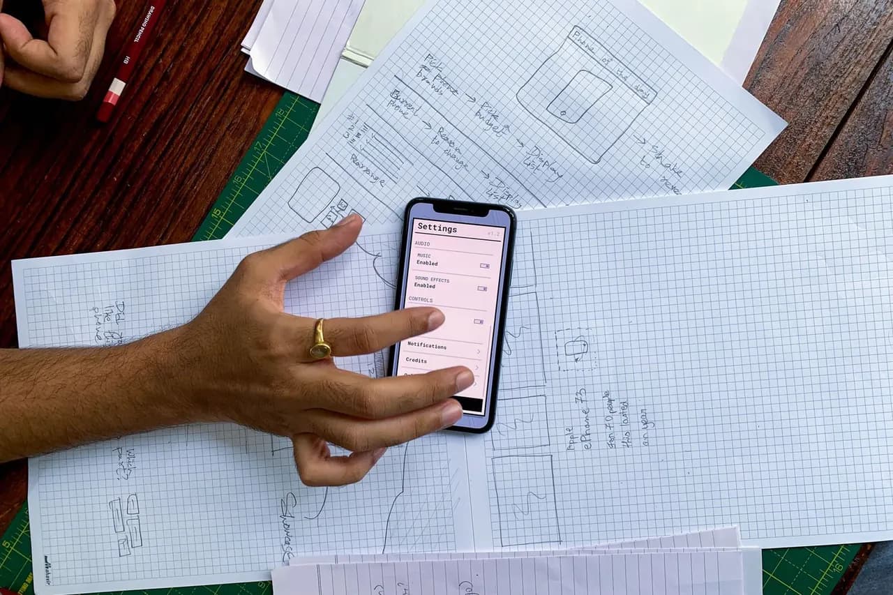 Designer reviewing a mobile app interface on a smartphone alongside hand-drawn wireframe sketches